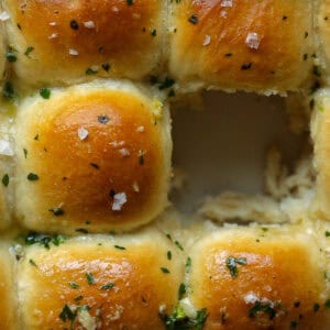 Close-up of golden-brown garlic vegan dinner rolls arranged in a grid pattern. Center roll is missing, revealing soft, fluffy interior. Sprinkled with herbs and salt, the bread features a shiny surface characteristic of melted butter.